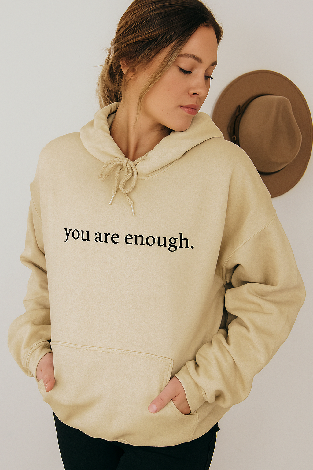 Woman wearing a beige hoodie with 'you are enough' text, standing against a plain background.